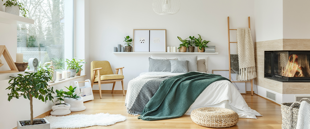 A cozy bedroom with large windows, a fireplace, and various plants. The room is decorated in a minimalist style with light colors and natural materials.